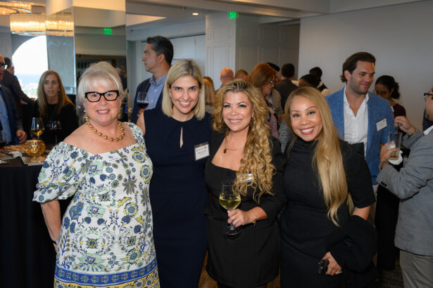 Four women pose and smile at a social event, holding drinks, with other people mingling in the background in a well-lit indoor space. South Florida Business & Wealth