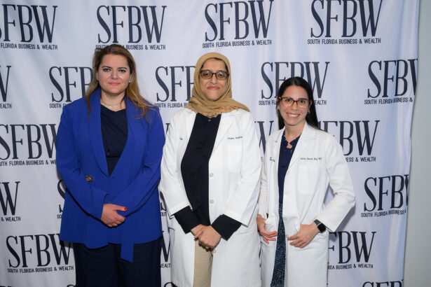 Three women stand side by side, two wearing white lab coats and one in a blue blazer, posing in front of a step-and-repeat banner with the SFBW (South Florida Business & Wealth) logo. South Florida Business & Wealth