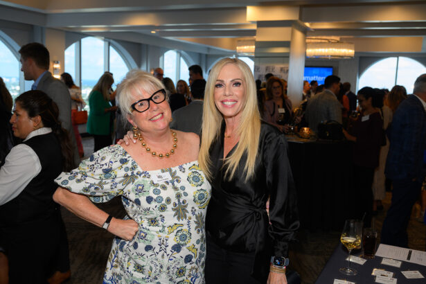 Two women smiling and posing together at a social event indoors, surrounded by other people. One woman wears glasses and a floral dress, the other has long blonde hair and wears a black blouse. Tables and large windows are in the background. South Florida Business & Wealth