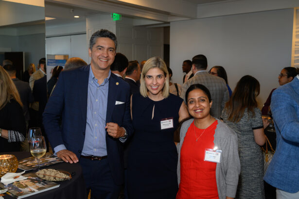 Three people, two women and one man, stand together at an indoor networking event, smiling at the camera. Other attendees mingle in the background. Name tags are visible, and there are drinks and food on tables nearby. South Florida Business & Wealth