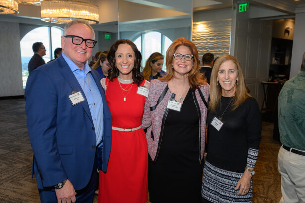 Four people stand together smiling at a professional event, each wearing nametags. The group includes one man in a blue suit and three women, all dressed in business attire, with a modern, well-lit room in the background. South Florida Business & Wealth