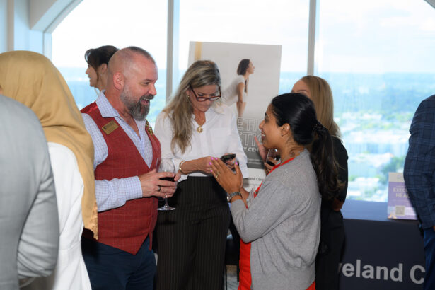 A group of people socialize at an indoor event. A man in a red vest holds a glass of wine, while a woman with glasses checks her phone. Others stand nearby, and a large window shows a city view in the background. South Florida Business & Wealth