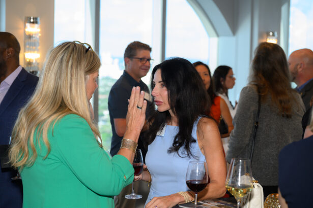 Two women stand and talk at a lively indoor social event; one wears a mint green jacket, the other a light blue dress. People mingle in the background, and glasses of wine are on the table in front of them. South Florida Business & Wealth