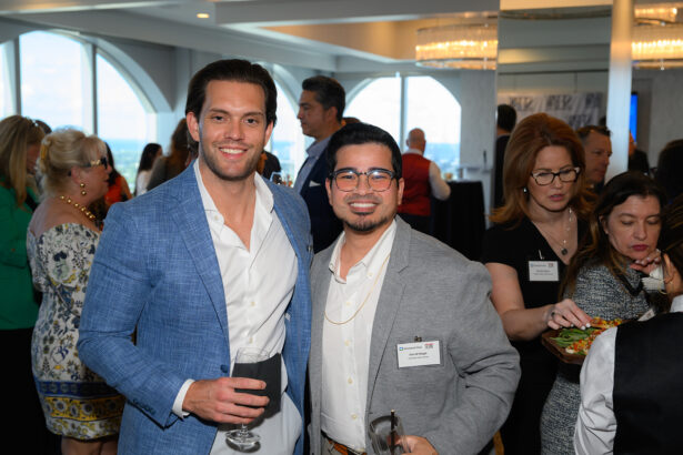 Two men dressed in business attire smile at the camera during a networking event. One holds a glass of wine. Other attendees are mingling and serving food in the background. The setting appears to be a bright, modern venue. South Florida Business & Wealth