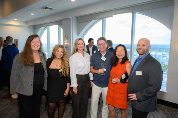 Six professionally dressed people, three men and three women, stand together smiling at an indoor event with large windows and a city view in the background. Some hold drinks and wear name tags. Other guests are visible behind them. South Florida Business & Wealth