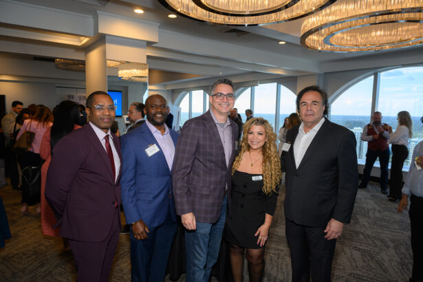 A group of five professionally dressed people pose and smile at a networking event in a modern, bright room with large windows and chandeliers. Other attendees are visible in the background. South Florida Business & Wealth