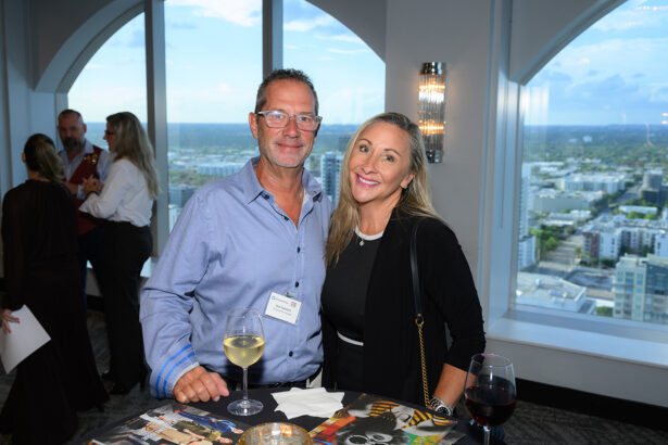 A man and woman pose and smile at a cocktail table with wine glasses, standing in front of large windows overlooking a cityscape at a social event. Other attendees are visible in the background. South Florida Business & Wealth