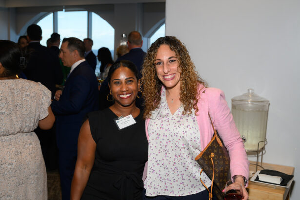 Two women smile at the camera at a social event. One wears a black dress, the other a pink blazer with a patterned shirt, holding a drink. People mingle in the background near large windows and a table with a glass beverage dispenser. South Florida Business & Wealth