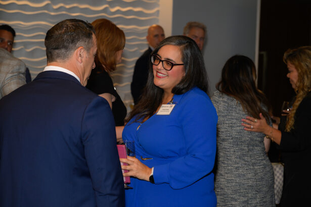 A woman in a bright blue dress and glasses smiles while talking to a man in a suit at a professional networking event. Other people are mingling in the background. South Florida Business & Wealth