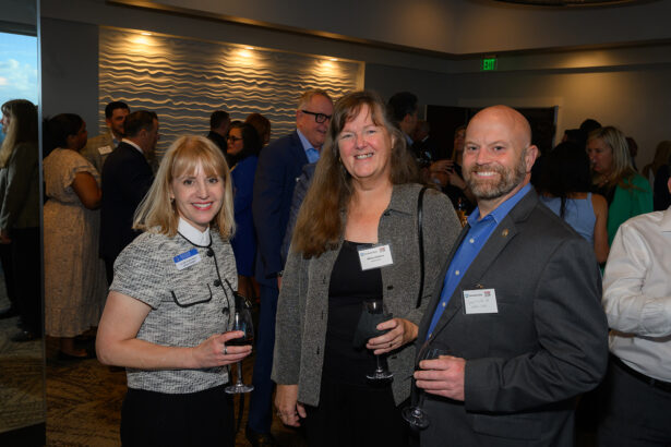Three people, two women and one man, stand indoors at a social event, smiling at the camera and holding glasses of wine. Other attendees mingle in the background under warm lighting. South Florida Business & Wealth