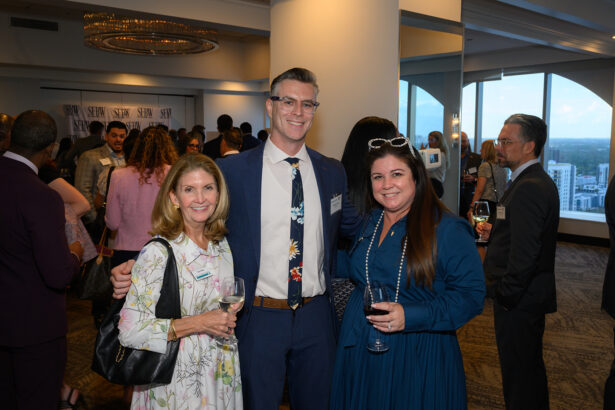 Three people dressed in business attire smile for a photo at a formal networking event. The room is filled with other attendees, and large windows reveal a cityscape in the background. South Florida Business & Wealth