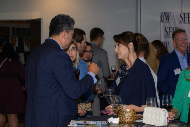 A man and woman in business attire converse at a networking event, holding drinks. Other people mingle in the background, and a table with wine glasses and decorative items is in the foreground. South Florida Business & Wealth