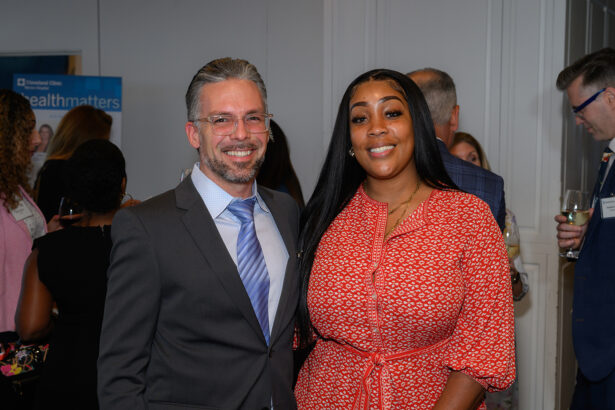 A man in a suit and a woman in a red patterned dress stand smiling at an indoor event, with other people mingling in the background. South Florida Business & Wealth