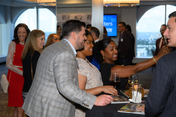A group of people dressed in business attire are smiling and talking around a high table at an indoor networking event. Some are holding drinks, and magazines and candles are on the table. Large windows show a city view. South Florida Business & Wealth