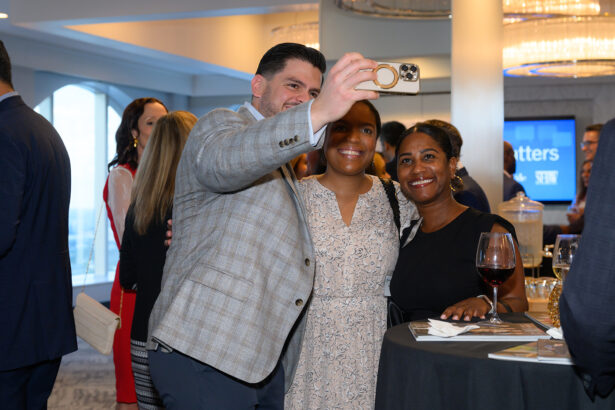 A man and two women smile and pose for a selfie at a social event, standing around a cocktail table with drinks and brochures. Other people mingle in the background. The atmosphere appears lively and friendly. South Florida Business & Wealth