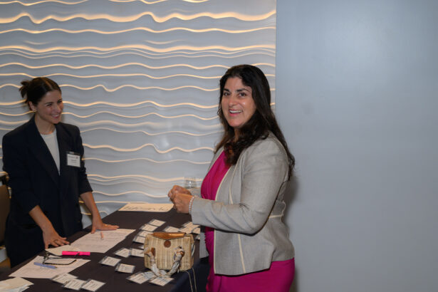 A woman in a gray blazer and pink dress smiles while standing at a registration table with name tags. Another woman behind the table is also smiling. The background features a wavy, textured wall. South Florida Business & Wealth
