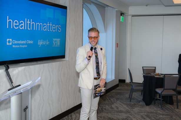 A man in a light suit and sunglasses holds a microphone while speaking at an indoor event. A screen behind him displays the text "healthmatters" and logos for Cleveland Clinic, Lifestyle, and SFH. South Florida Business & Wealth
