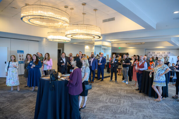 A group of professionally dressed people stand and listen attentively in a well-lit event room with large chandeliers, high ceilings, and networking tables. South Florida Business & Wealth