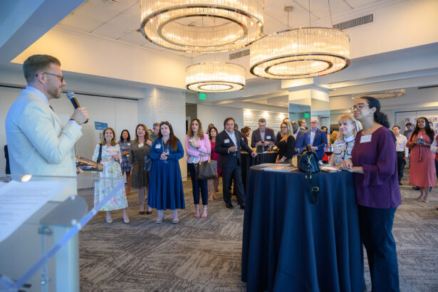 A man in a light suit speaks into a microphone to a group of people in business attire who are listening attentively at a formal indoor event with modern chandeliers. South Florida Business & Wealth