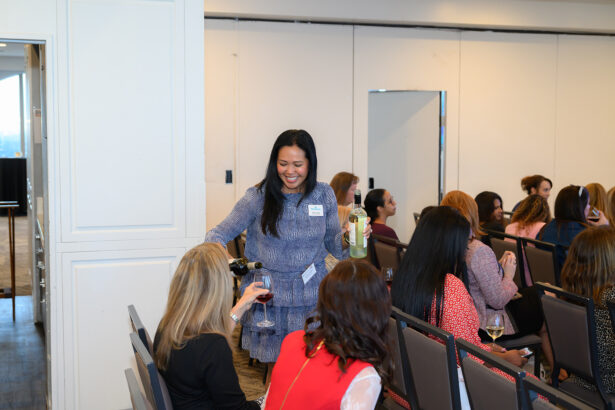 A woman in a blue dress smiles as she pours wine for seated attendees at an indoor event. Other people are seated in rows, some conversing, in a brightly lit room. South Florida Business & Wealth