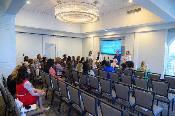 A group of people sits in rows of chairs facing a panel of speakers at the front of a bright conference room. A large screen behind the speakers displays “healthmatters.”. South Florida Business & Wealth