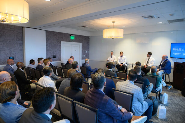 A group of people in business attire attend a panel discussion in a conference room. Four speakers sit at the front while the audience listens attentively, facing them. South Florida Business & Wealth