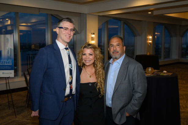 Three people in business attire stand together and smile at an indoor event with large windows and nighttime city views in the background. Two men are on either side of a woman with long curly hair. South Florida Business & Wealth