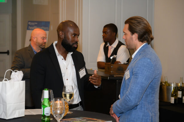 Two men in business attire are having a conversation at a social event. Drinks and gift bags are on the table. Other people are in the background, including a bartender and another attendee. South Florida Business & Wealth