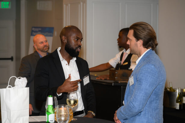 Two men in business attire are talking at a networking event. One gestures while speaking; drinks and gift bags are on the table. Other people are in the background near a counter. The setting appears professional and social. South Florida Business & Wealth