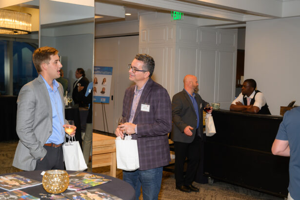 Two men in business attire converse while holding drinks and gift bags at a networking event. In the background, two other men chat near a bar where a bartender stands behind the counter. The setting is modern and well-lit. South Florida Business & Wealth