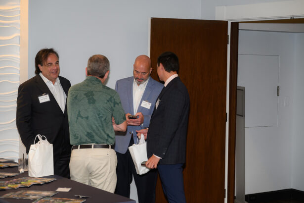 Four men in business attire stand together near a door, engaged in conversation. Two are holding white gift bags. A table with magazines or brochures is visible in the foreground. South Florida Business & Wealth