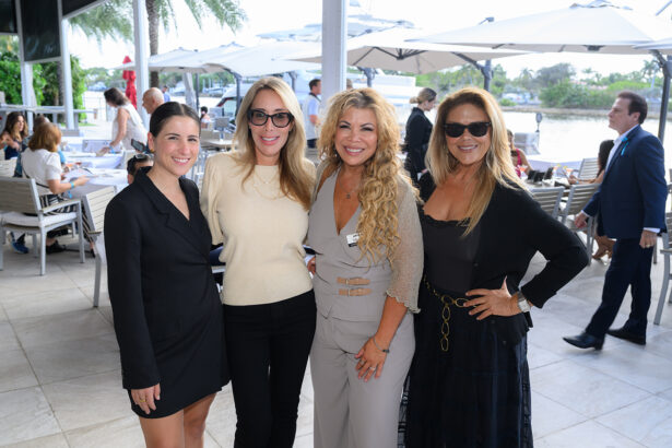 Four women stand together smiling at an outdoor patio restaurant with tables, umbrellas, and other people in the background. Palm trees and water are visible behind them on a bright day. South Florida Business & Wealth