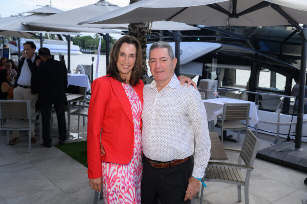 A smiling man and woman pose together outdoors by a marina, with yachts, tables, chairs, and umbrellas in the background. The woman is wearing a red jacket and patterned dress, and the man is in a white shirt and dark pants. South Florida Business & Wealth