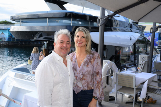 A smiling man and woman stand together on a dock beside a luxury yacht. The man wears a white shirt and the woman a light purple blouse. Outdoor tables and umbrellas are set up nearby, with water and other people in the background. South Florida Business & Wealth