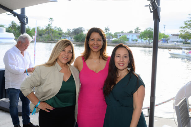 Three women smile and pose together outdoors by a waterfront, with boats and trees in the background. One woman wears green, another pink, and the third dark green. A man stands in the background near the water. South Florida Business & Wealth