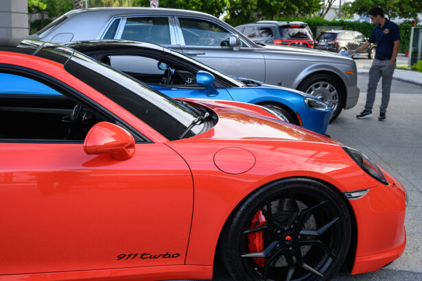 Three luxury cars—a red Porsche 911 Turbo, a blue sports car, and a silver Rolls-Royce—are parked in a row outside, with a man standing nearby on the sidewalk. South Florida Business & Wealth
