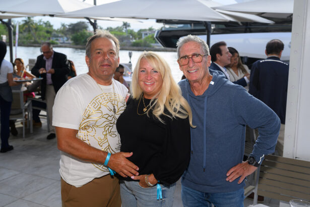 Three adults stand close together and smile at an outdoor gathering near water, with tables, people, and white umbrellas visible in the background. South Florida Business & Wealth
