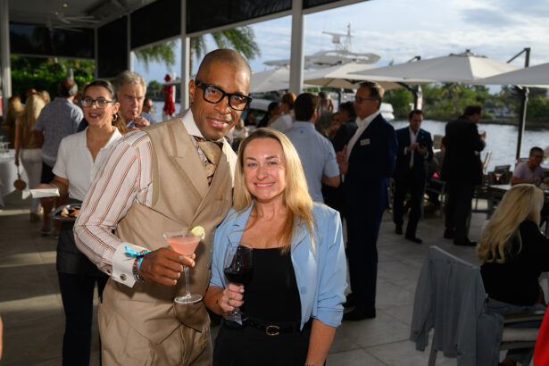 A man in a beige suit and a woman in a light blue blazer smile and pose with drinks at an outdoor social gathering near a waterfront, with other guests and yachts in the background. South Florida Business & Wealth