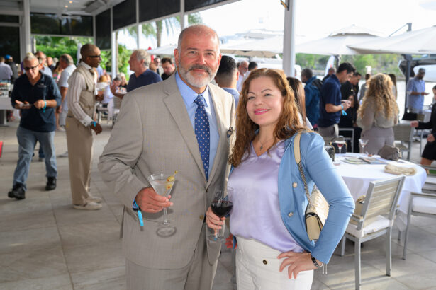 A man in a light gray suit holding a martini and a woman in a blue jacket holding red wine pose and smile at an outdoor social event with people mingling in the background. South Florida Business & Wealth