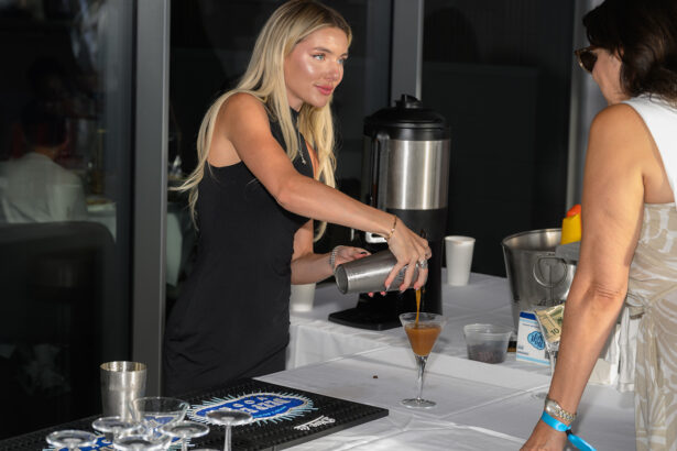 A woman with long blonde hair in a black dress pours a drink from a shaker into a martini glass at a bar setup, while another woman in a white dress stands nearby. The table holds various bar tools and supplies. South Florida Business & Wealth