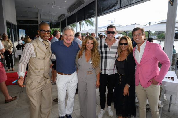 Six people stand closely together and smile for a group photo at an indoor-outdoor event space, with other attendees and tables visible in the background. The group is dressed in smart casual to semi-formal attire. South Florida Business & Wealth