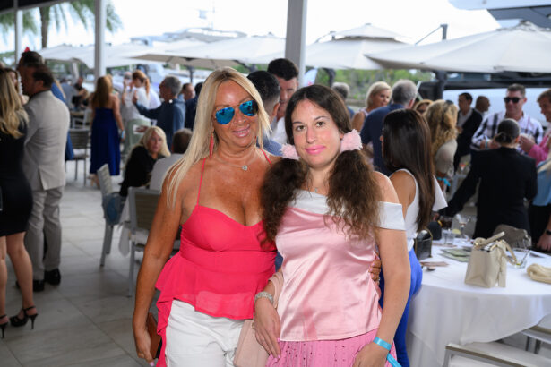 Two women pose together at an outdoor social event. The woman on the left wears sunglasses and a bright pink top, while the woman on the right wears a pink and white outfit. People mingle in the background under umbrellas. South Florida Business & Wealth
