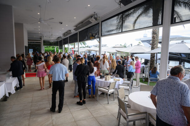 A large group of people socialize at an outdoor event space with tables and umbrellas. Guests are dressed in semi-formal attire, and the setting is bright with palm trees visible outside. South Florida Business & Wealth