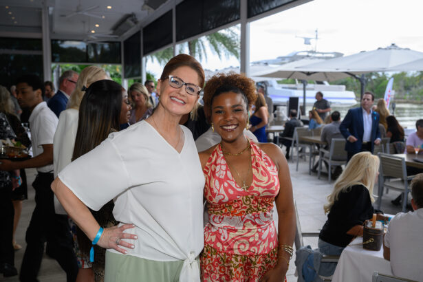 Two women smiling and posing together at an outdoor social event; people are gathered and mingling in the background near tables, with palm trees and a docked boat visible outside. South Florida Business & Wealth