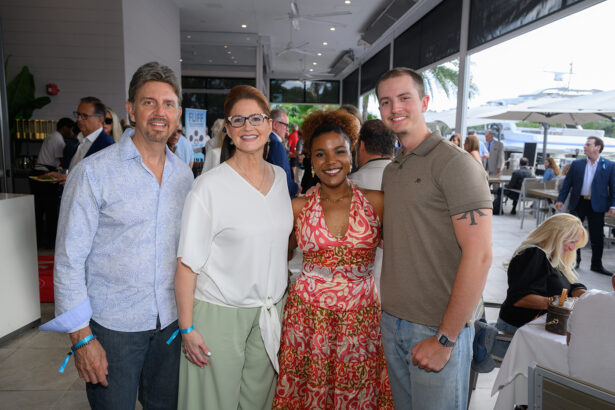 Four people stand smiling together indoors at a casual event, two men and two women, with other people mingling and sitting at tables in the background near large windows and outdoor patio umbrellas. South Florida Business & Wealth