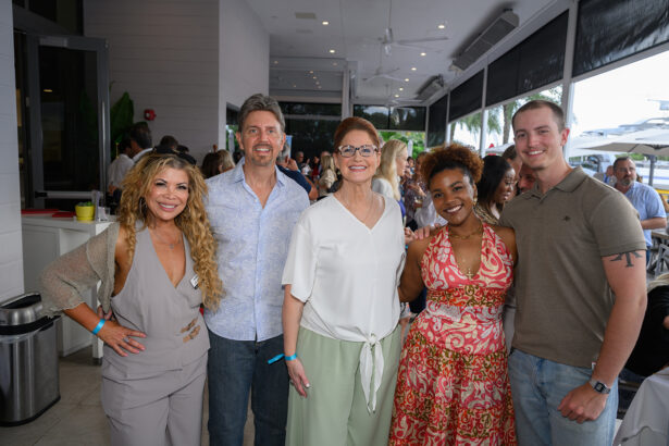 Five people smile and pose together indoors at a social event. There are tables and other guests in the background, and large windows show greenery and outdoor seating. Everyone looks happy and relaxed. South Florida Business & Wealth