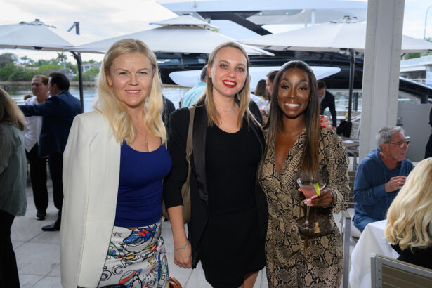 Three women stand close together, smiling at an outdoor event. They are dressed in stylish outfits, and one holds a glass of wine. Other people are seated and mingling in the background near yachts and umbrellas. South Florida Business & Wealth