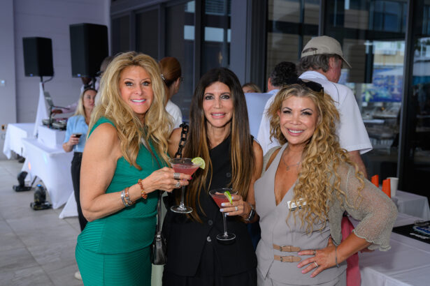Three women dressed in stylish outfits smile at the camera while holding cocktails at an indoor-outdoor social event. Other people and event tables appear in the background. South Florida Business & Wealth
