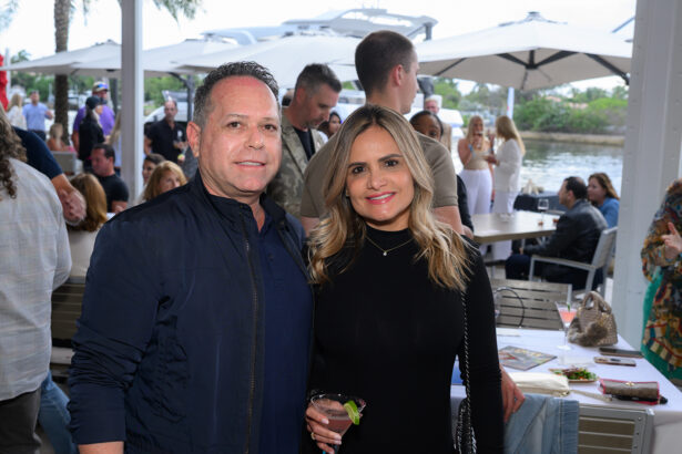 A man in a navy jacket and a woman in a black dress smile at an outdoor restaurant by the water, surrounded by other people dining and socializing under large white umbrellas. South Florida Business & Wealth
