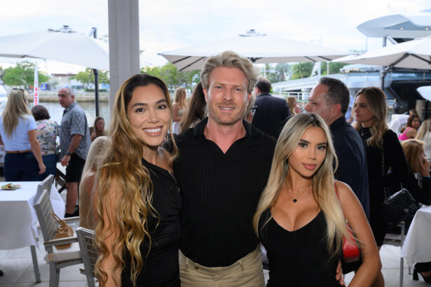 Three people pose and smile for a photo at an outdoor event with white tables, umbrellas, and other guests in the background. The group is dressed in black attire and looks cheerful. South Florida Business & Wealth